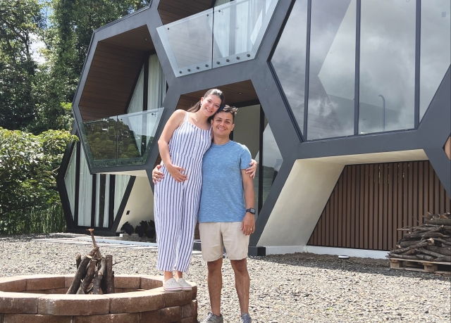 Esteban Arguedas, wearing a blue t-shirt and tan shorts, stands next to his wife Marie Jesus, wearing a white and blue striped romper, who stands on the edge of a brick fire pit. Behind them is a metal and glass house designed out of hexagons.