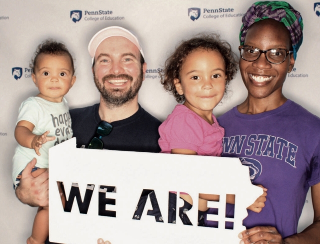 Karen Kemirembe, wearing a purple t-shirt with the words "Penn State" in navy blue, stands and holds a white sign with cut out letters that say "We Are!" Standing next to her is her husband and her two young daughters.