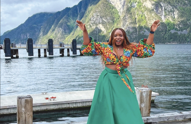 Nicole Magabo, wearing a rainbow patterned blouse and teal skirt, holds her hands up in the air and smiles while standing on a dock in front of a lake and mountain.