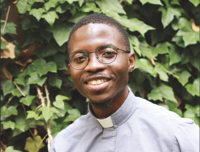 Pastor Menzi Nkambule, wearing a grey neckband shirt, smiles for a headshot in front of a leafy, green, background.