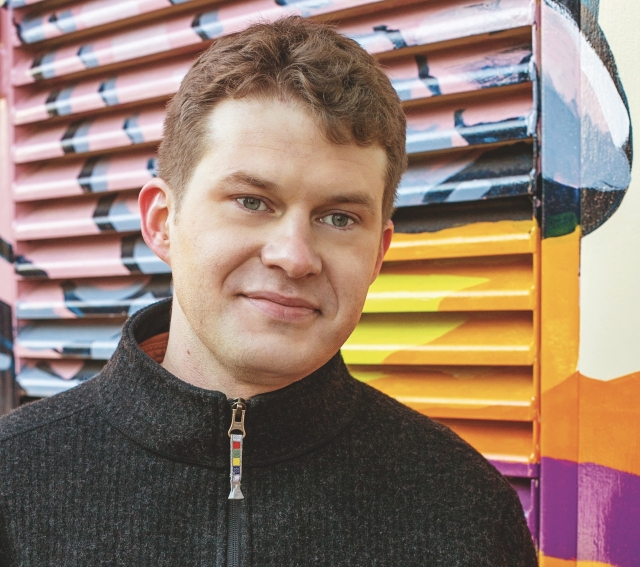 Peter Sutoris, wearing a dark gray quarter-zip, poses for a headshot in front of a bright and multi-colored background.