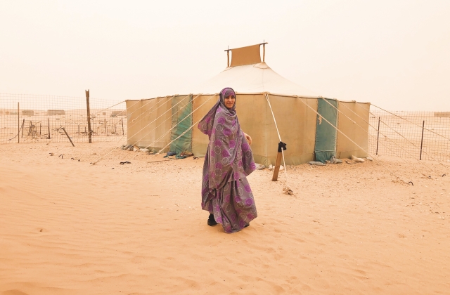 Senia Bachir-Abderahman, wearing a gray and purple patterned abaya, stands and faces the camera in front of a tent put up in a vast desert.