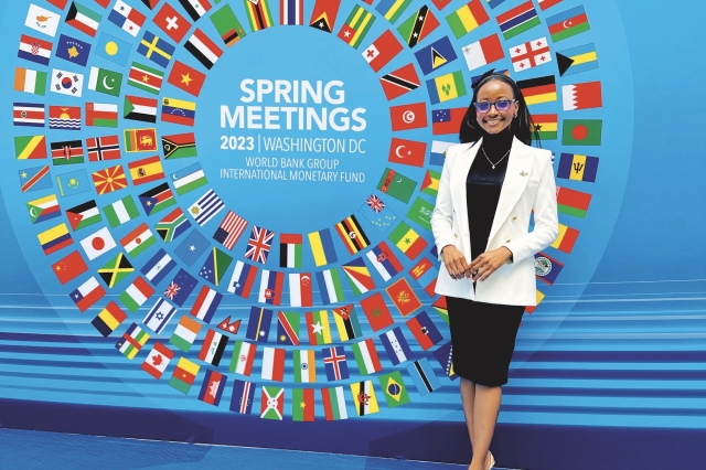 Tatenda Dzvimbo, wearing a white and black pantsuit, poses for a photo next to a mural featuring a swirling circle of global flags on a blue background, and the text "Spring Meetings 2023 Washington D.C."