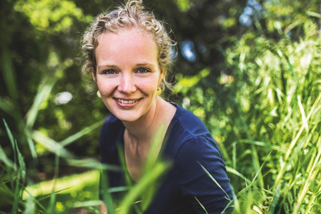 Nina Overgaard Therkildsen, wearing a blue shirt, crouches amidst tall, green, grass for a portrait.