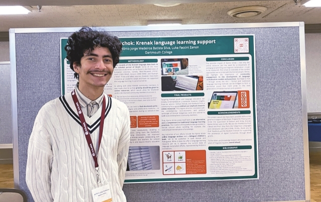 Antonio Jorge Medeiros Batista Silva, wearing a white sweater over a white button-down, smiles posing for a photo in front of a colorful academic poster pinned up to a bulletin board.