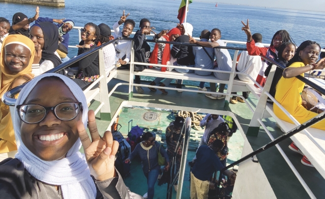 Anna Diagne Sène, wearing a white hijab and a black shirt, flashes the peace scene while taking a selfie in front of a large group of young adults on the top deck of a boat out on the water.