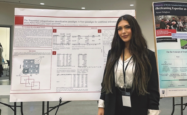 Aylin Ak, wearing a black blazer and white blouse, poses for a photo in front of a data-heavy academic poster mounted to a tri-pod in a conference room with other posters.