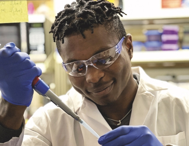 Melvin Osei Opoku, wearing a white lab coat, using an electric pipette to put liquid in a vial. Behind him are shelves of supplies in a research laboratory.
