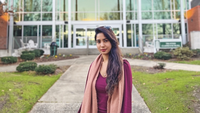 Zakia Maryam Najafizada, wearing a maroon shirt and a large pink scarf, poses for a portrait in front of building made of glass and white metal.