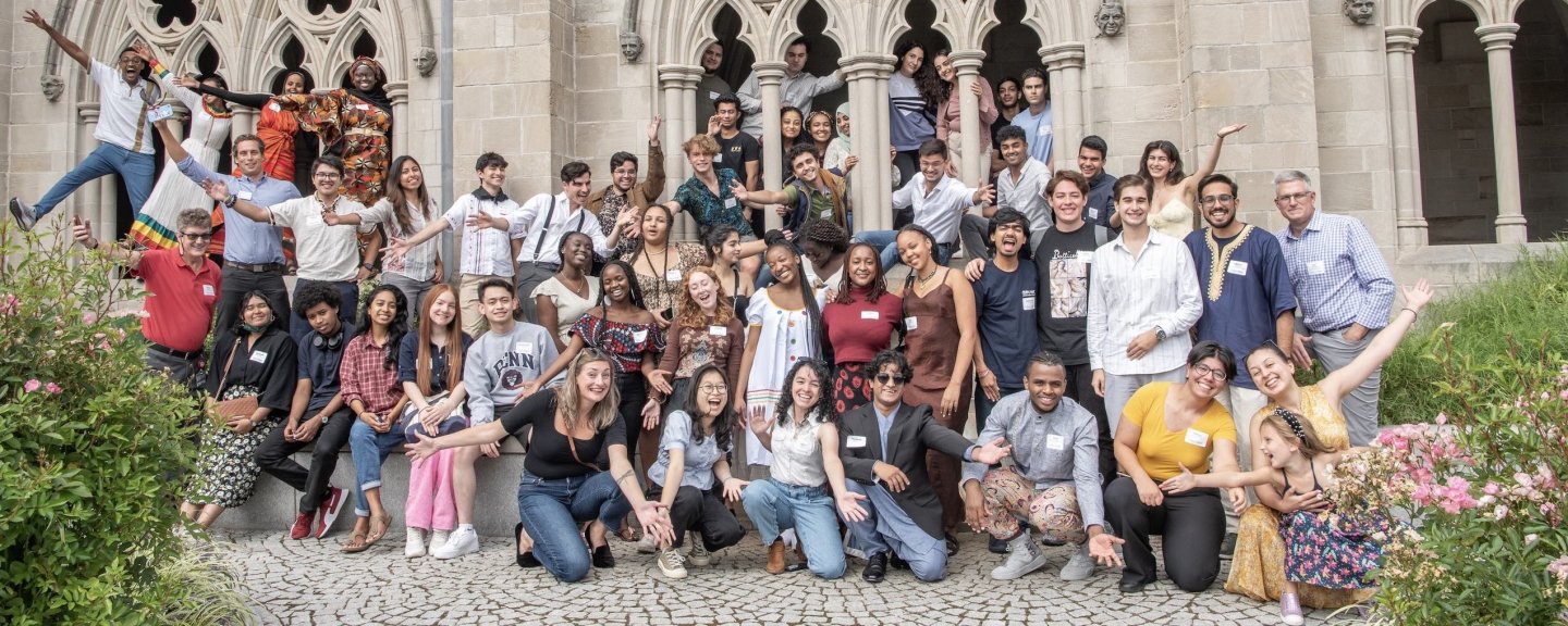 A group of college students of varying ages, heights, skin colors, and clothing colors pose in various smiling and goofy ways on and around the side of a stone building.