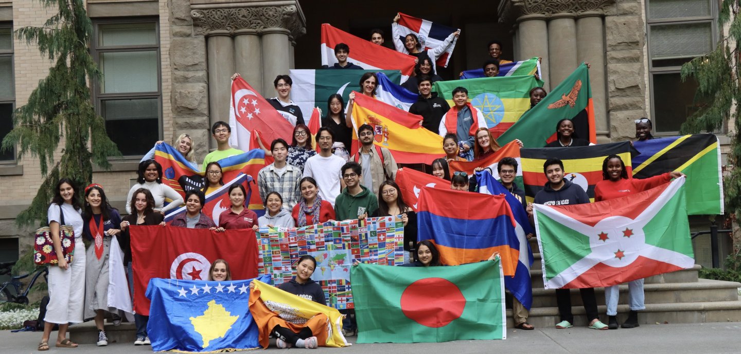 A group of college students of various skin tones and appearances pose together on stone steps holding an array of world flags.