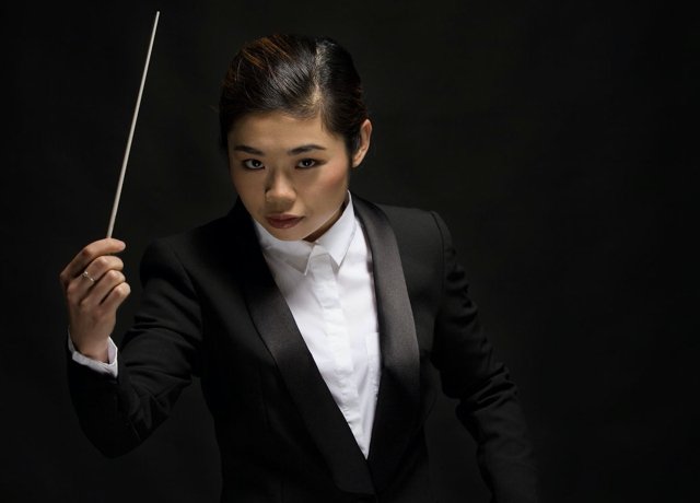 Elim Chan, wearing a classical black and white tuxedo, gestures with a conductor's baton in front of a black background.