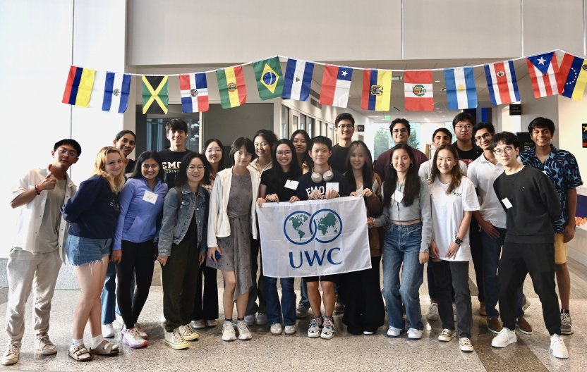 A group of college students pose together smiling, and the two in the center hold a white flag with the blue text UWC 